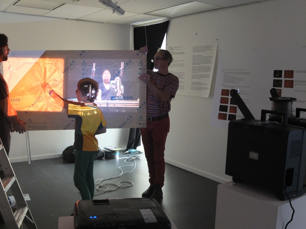 An image of a young boy standing in front of a projection screen. The image on the screen shows artist Lynn Loo performing her work Autumn Fog. On the wall is the score to create the performance for this work.
